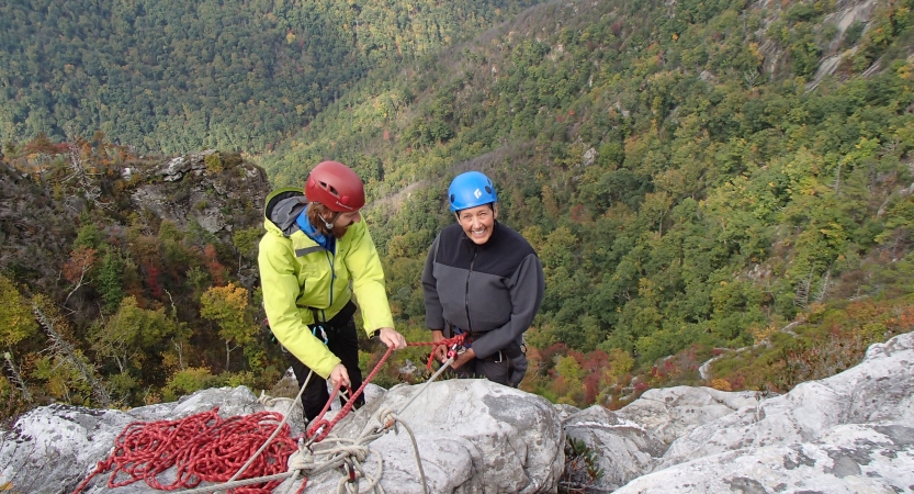 Two people wearing safety gear are secured by ropes near the edge of a cliff. One person appears to be an instructor, giving direction to the other person.